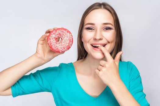 Portrait of cheerful young beautiful girl in blue blouse standing, holding and showing pink donut, sucking her finger with toothy smile in grey background. Indoor, isolated, copy space, studio shot
