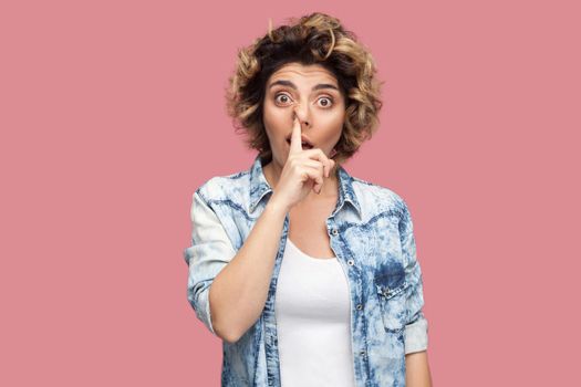 Portrait of crazy foolish young woman with curly hairstyle in casual blue shirt standing with finger on her nose, drilling and looking at camera. indoor studio shot, isolated on pink background.