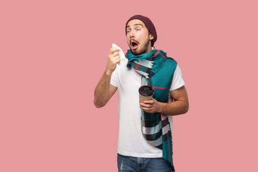Sickness modern bearded young hipster man in white shirt, hat and scarf standing, sneezes on napkin with opened mouth, holding hot tea in paper cup. Indoor, isolated,studio shot, pink background
