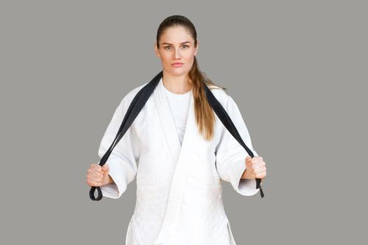 Confident attractive athletic woman in white kimono standing and wraps black belt around the neck, looking at camera. Japanese karate and sports concept. Indoor, studio shot, isolated, grey background