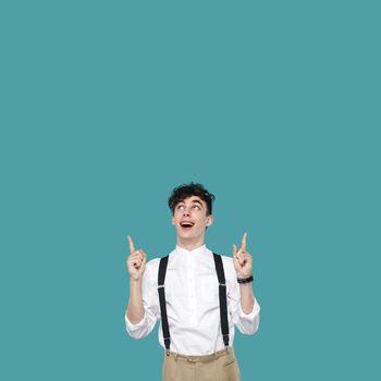 Surprised man pointing and looking at top copyspace. Portrait of handsome hipster curly young businessman in classic casual white shirt and suspender standing. studio shot isolated on blue background.