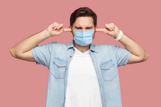 I don't want to hear you. Portrait of confused young man with surgical medical mask in blue shirt standing and putting fingers on ears and dont care. indoor studio shot, isolated on pink background
