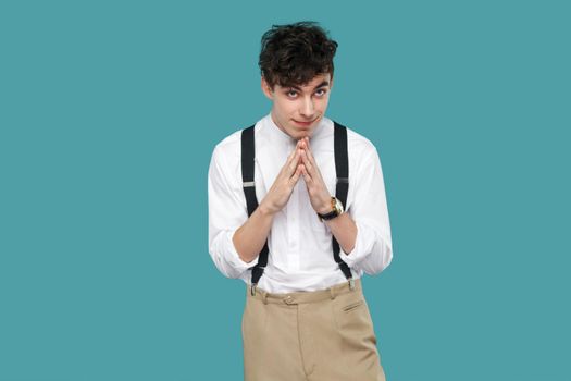 cunning funny man with plan looking at camera. Portrait of handsome hipster curly young businessman in classic casual white shirt and suspender standing. indoor studio shot isolated on blue background