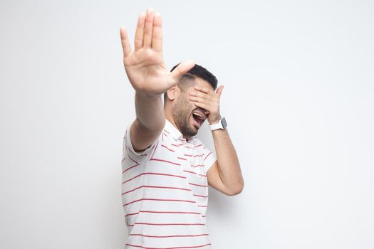 Stop, i don't want to look at this. Portrait of confused bearded young man in striped t-shirt standing, covering his eyes and showing stop gesture. indoor studio shot, isolated on white background.