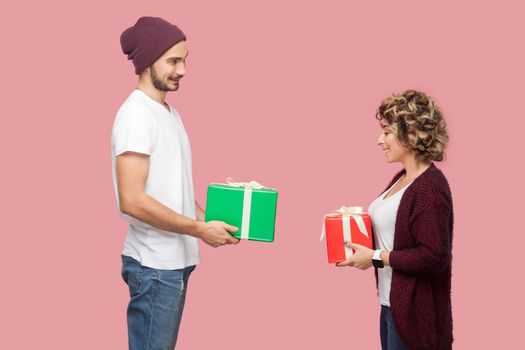 Side view portrait of suprised couple of friends in casual style standing, giving present boxes to each other, celebrate anniversary, toothy smile. Isolated, indoor, studio shot, pink background