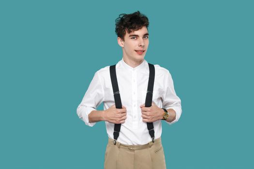 happy man toothy smile, standing and looking at camera. Portrait of handsome hipster curly young man in classic casual white shirt and suspender. indoor studio shot isolated on blue background.