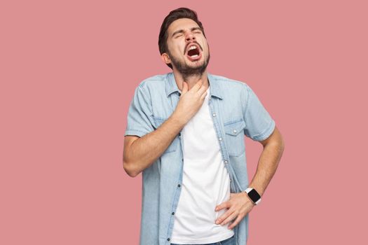 Throat pain or cold flu. Portrait of sad sick handsome bearded young man in blue casual style shirt standing and holding his painful neck and screaming. indoor studio shot, isolated on pink background