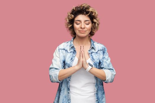 Portrait of calm young woman with curly hairstyle in casual blue shirt standing with closed eyes, palm hands, smile and doing yoga meditating. indoor studio shot, isolated on pink background.