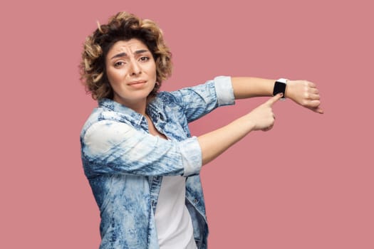 I need more time. Portrait of young pleased woman with curly hairstyle in casual blue shirt standing and pointing at her smart watch and want more time. indoor studio shot, isolated on pink background