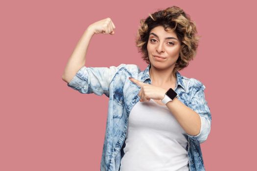 Portrait of strong proud young woman with curly hairstyle in casual blue shirt standing and pointing at her biceps to show her independent or feminism. indoor studio shot, isolated on pink background.