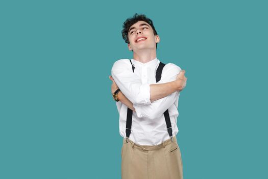 Happy excited man toothy smile and hug himself. Portrait of handsome hipster curly young businessman in classic casual white shirt, suspender standing. indoor studio shot isolated on blue background