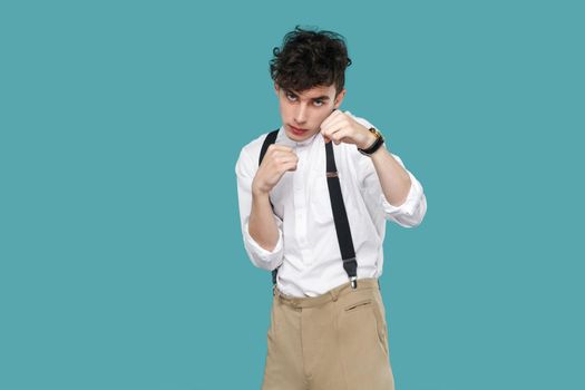 Serious man with boxing fist looking at camera. Portrait of handsome hipster curly young businessman in classic casual white shirt, suspender standing. indoor studio shot isolated on blue background.