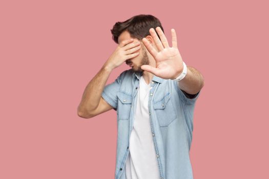 Stop, I don't want to see this. Portrait of bearded young man in blue casual style shirt standing, covering his eyes and showing stop hand sign gesture. indoor studio shot, isolated on pink background