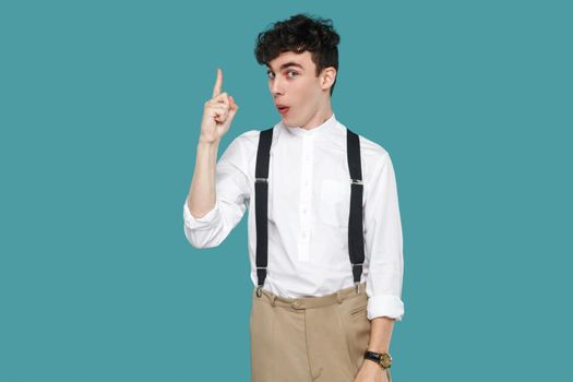 Amazed suprised man with idea looking at camera. Portrait of handsome hipster curly young businessman in classic casual white shirt, suspender standing. indoor studio shot isolated on blue background.