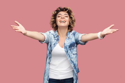 Give me hug. Portrait of happy young woman with curly hair in casual blue shirt standing with raised arms to embrace and looking at camera, toothy smile. indoor studio shot isolated on pink background