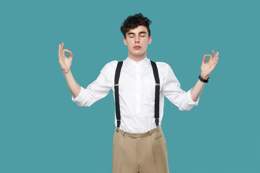 Calm man raised arms in yoga hands and closed eyes. Portrait of handsome hipster curly young businessman in classic casual white shirt and suspender standing. studio shot isolated on blue background.