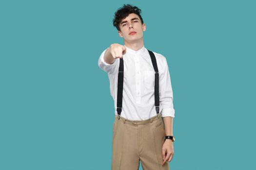 Serious man pointing and looking at camera. Portrait of handsome hipster curly young businessman in classic casual white shirt and suspender standing. indoor studio shot isolated on blue background.