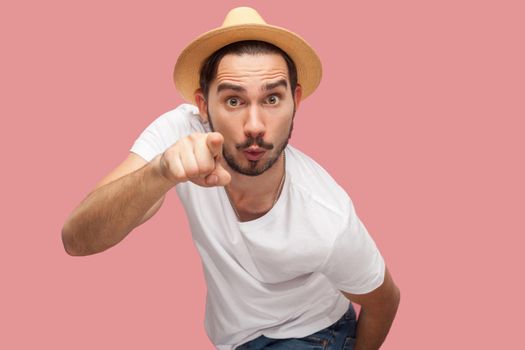 Portrait of shocked bearded young man in white shirt with hat standing, looking and pointing at camera with surprised face. indoor studio shot, isolated on pink background copyspace.