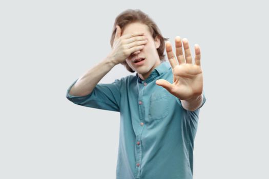 Stop, I don't to see this. Portrait of handsome long haired blonde young man in blue casual shirt standing with block hand and covering his eyes. indoor studio shot, isolated on light grey background.