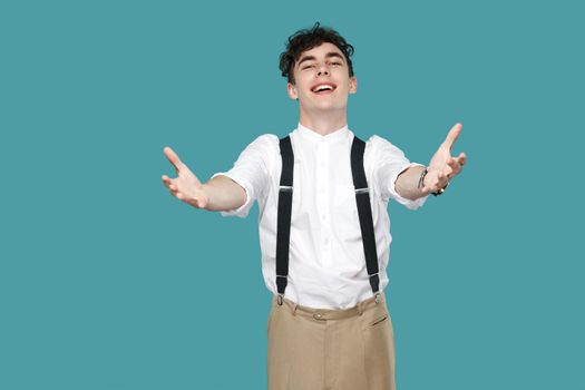 Happy man toothy smile at camera and giving hands to hug. Portrait of handsome hipster curly young businessman in classic casual white shirt and suspender. studio shot isolated on blue background.