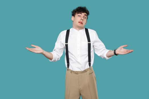 I don't know. confused man raised arms and looking at camera. Portrait of handsome hipster curly young businessman in classic casual white shirt and suspender. studio shot isolated on blue background.