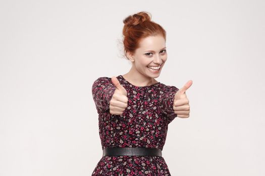 Happy cheerful redhead woman showing thumb up and toothy smile. Isolated studio shot on gray background
