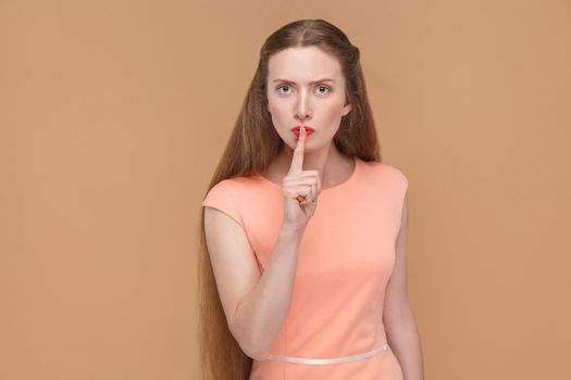 shhh sign, serious woman touching her lips with finger. portrait of emotional cute, beautiful woman with makeup and long hair in pink dress. studio shot, isolated on light brown or beige background.