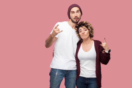 Portrait of funny couple of friends in casual style standing, hugging each other and showing rock and roll hand sign gesture, looking at camera. Isolated, indoor, studio shot, pink background