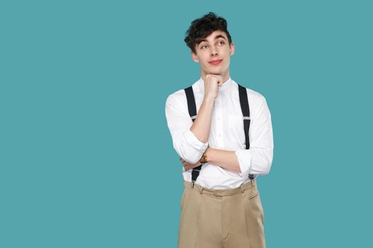 Dreamy man standing, dreaming and looking away. Portrait of handsome hipster curly young businessman in classic casual white shirt, suspender standing. indoor studio shot isolated on blue background.