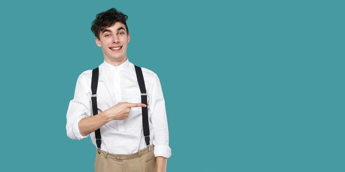 Happy man pointing at empty copyspace and looking at camera with toothy smile. Portrait of hipster curly young businessman in classic casual white shirt suspender. studio shot isolated blue background