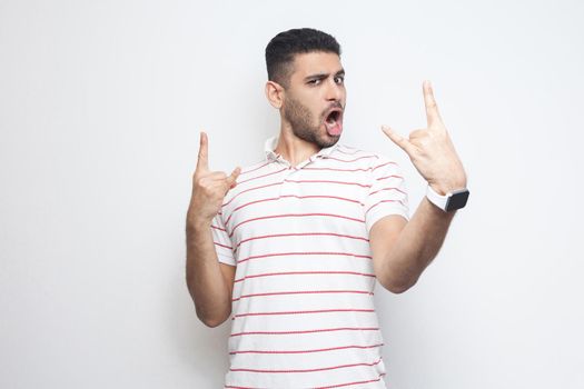 Portrait of funny handsome bearded young man in striped t-shirt standing with rock and roll horns gesture and looking at camaera with surprised face. indoor studio shot, isolated on white background.