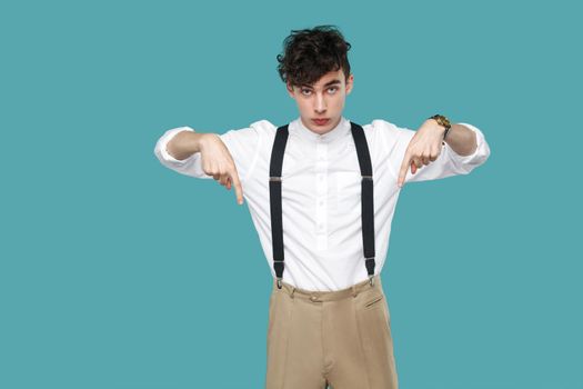 Here and right know. serious man pointing down and looking at camera. Portrait of handsome hipster curly young businessman in classic white shirt, suspender. studio shot isolated on blue background.