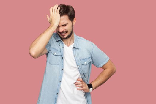 Portrait of sad alone loser bearded young man in blue casual style shirt standing holding his head down and thinking what to do. indoor studio shot, isolated on pink background.