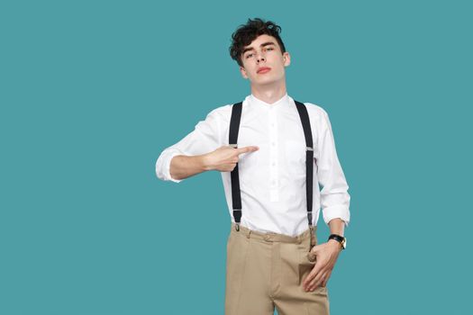 This is me. proud man pointing himself and looking at camera. Portrait of handsome hipster curly young businessman in classic casual white shirt and suspender. studio shot isolated on blue background.