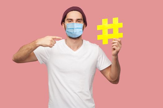 Portrait of happy young hipster man with surgical medical mask in white shirt and casual hat standing, holding yellow hashtag and pointing finger to it. Indoor, isolated, studio shot, pink background