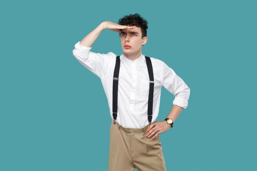Attentive man looking to far with hand on forrhead. Portrait of handsome hipster curly young businessman in classic casual white shirt and suspender standing. studio shot isolated on blue background.
