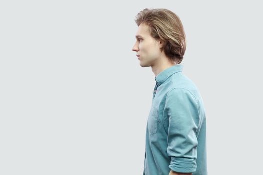 Profile side view portrait of handsome long haired blonde young man in blue casual shirt standing and looking with serious face. indoor studio shot, isolated on light grey background.