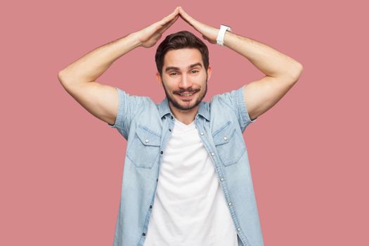 Portrait of happy satisfied handsome bearded young man in blue casual shirt standing with roof gesture hands on head and looking at camera with toothy smile. studio shot, isolated on pink background.