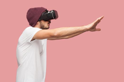 Side view of portrait of wondering bearded young hipster man in white shirt and casual hat standing, wearing vr playing video game with raised arms. Indoor, isolated,studio shot, pink background