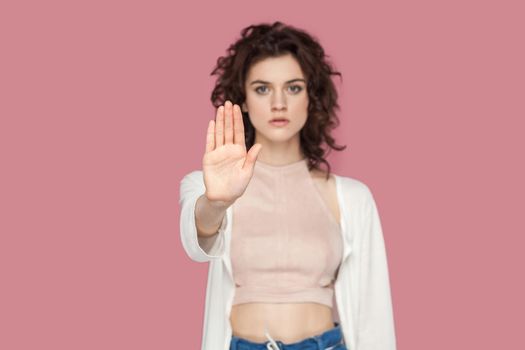 Stop. Portrait of serious beautiful brunette young woman with curly hairstyle in casual style standing with stop hands gesture and caution to dont touch. indoor studio shot isolated on pink background