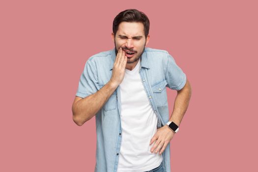 toothache or pain. Portrait of sad sick bearded young man in blue casual style shirt standing and touching his chik because feeling pain on tooth. indoor studio shot, isolated on pink background.
