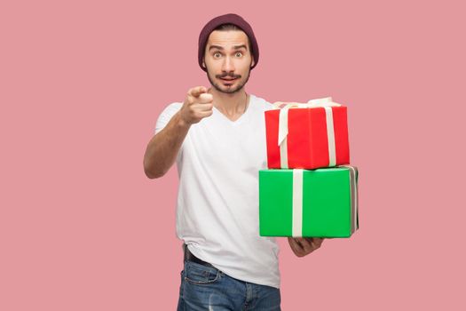 Portrait of suprised handsome bearded young hipster man in white shirt and casual hat standing, holding two present box, pointing finger to camera. Indoor, isolated, studio shot, pink background