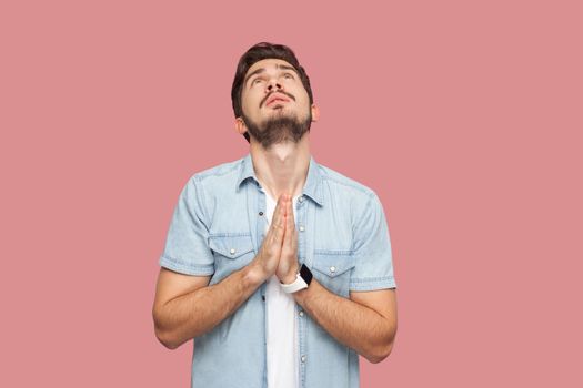 My God please help me. Portrait of hopeful sad handsome bearded young man in blue casual style shirt standing looking up and praying with worry face. indoor studio shot, isolated on pink background.