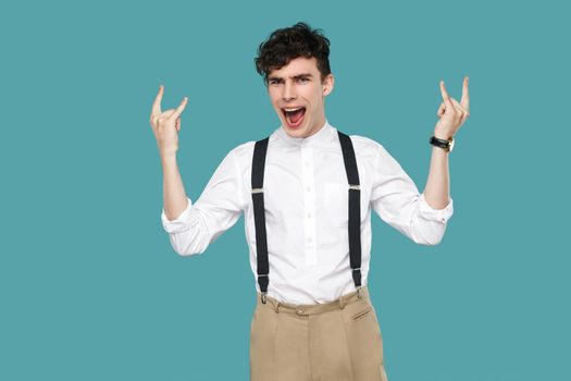 Man screaming and showing rock sign gesture. Portrait of handsome hipster curly young businessman in classic casual white shirt and suspender standing. indoor studio shot isolated on blue background.