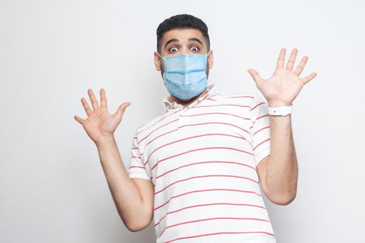 Portrait of shocked young with surgical medical mask man in striped t-shirt standing with raised arms and looking at camera with big eyes and panic. indoor studio shot, isolated on white background.