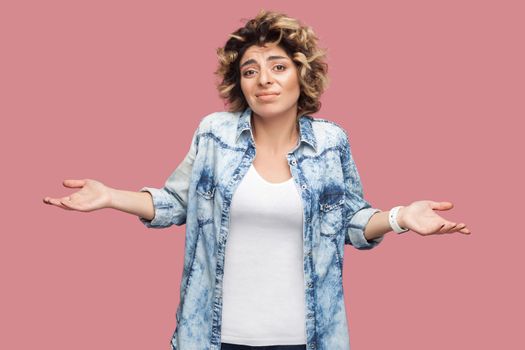 I don't know. Portrait of confused young woman with curly hairstyle in casual blue shirt standing with raised arms and looking at camera and thinking. indoor studio shot, isolated on pink background.