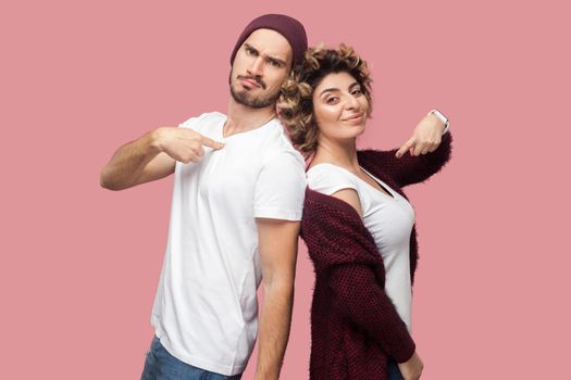 Portrait of satisfied successful couple of friends in casual style standing back to back, pointing themself with proud face and looking at camera. Isolated, indoor, studio shot, pink background