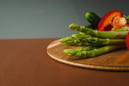 Vegetables lie on a wooden board: tomatoes, asparagus, cucumbers, red bell peppers. brown, dark gray background. place for text