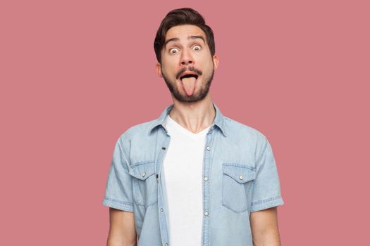 Portrait of funny crazy handsome bearded young man in blue casual style shirt standing with big eyes, tongue out and looking at camera. indoor studio shot, isolated on pink background.