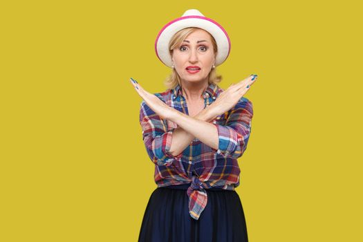 Stop. There is no way. Portrait of serious modern stylish mature woman in casual style with hat standing with X sign and looking at camera. indoor studio shot isolated on yellow background.